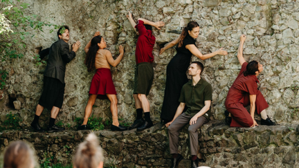 Ein Gruppe von Menschen bewegt sich an einer Steinmauer entlang; ein Junger Mann sitzt davor