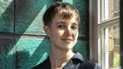 Junge Frau vor einer Wand. Sie schaut in die Kamera. Es ist die Autorin Tara Meister.