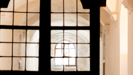 Blick durch eine dunkle, verglaste Tür in einen lichtdurchfluteten historischen Kreuzgang der Kartause Mauerbach. Helle Gewölbe, barocke Architektur und sanftes Tageslicht prägen die stimmungsvolle Atmosphäre.