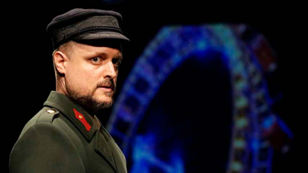 Schauspieler in historischer Militäruniform mit Mütze vor blau beleuchtetem Riesenrad auf einer Bühne