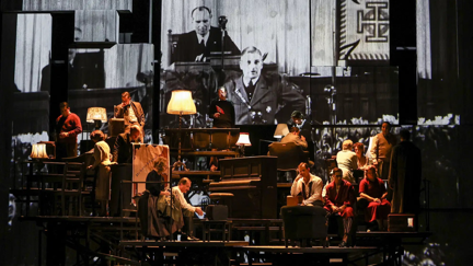 Große Theaterszene von dem Stück Lass uns die Welt vergessen - Volksoper 1938 mit mehreren Darstellenden vor einer projizierten Schwarz-Weiß-Filmaufnahme im Hintergrund.